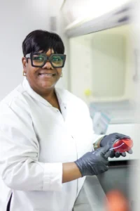 Food safety technician in a white lab coat and gloves holding a petri dish in a laboratory setting.